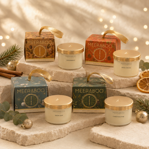 Six Meeraboo holiday candles in glass jars with gold lids are arranged on stone platters with their branded gift boxes. The set includes Christmas Pine, Snow Gum, Spiced Orange and Gingerbread styled with eucalyptus, cinnamon, dried orange slices, and gold ornaments against a warm, softly lit background.