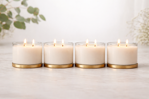 Four lit candles with gold bases on a white surface, surrounded by greenery.