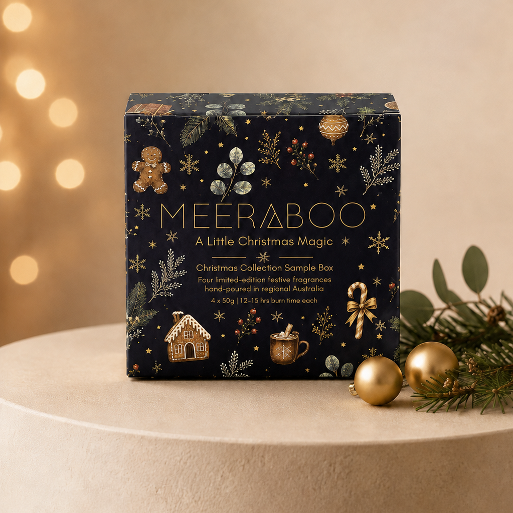 A MEERABoo 'A Little Christmas Magic' Christmas collector's sample box, decorated with gold and silver festive illustrations on a black background, displayed on a beige pedestal with holiday lights and ornaments in the background.