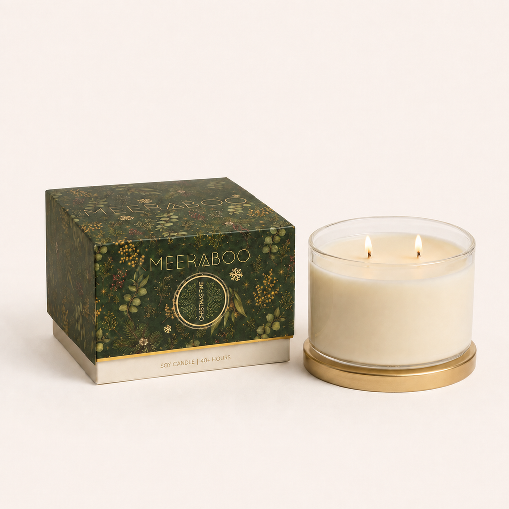 A lit two-wick soy candle in a clear glass jar with a gold base, shown next to its illustrated green and gold box. The box is branded MEERA BOO.