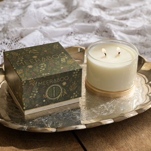 A lit Christmas candle in a clear glass jar sits on a silver tray beside its decorative green and gold MEERABOO box, placed on a wooden table with white lace fabric in the background.