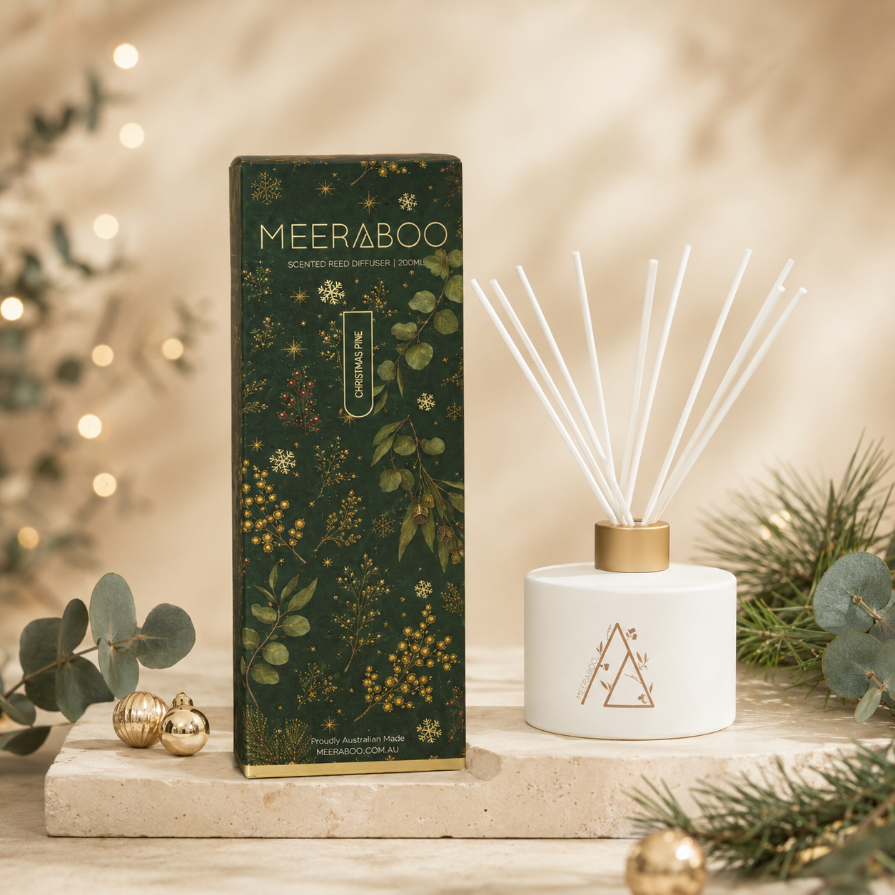 Four Meeraboo reed diffuser boxes in festive holiday packaging are arranged on stone blocks with a white reed diffuser bottle and reeds in front. The boxes are dark green, teal, rust red, and warm brown with gold-toned holiday illustrations, surrounded by pine branches, gold ornaments, and warm string lights.