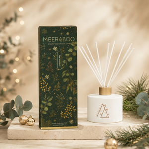 A reed diffuser and its decorative box displayed on a stone slab with holiday greenery and warm bokeh lights in the background. The box is dark green with botanical illustrations and the brand name MEERABOO visible, and the diffuser has white reeds in a white ceramic bottle with a gold cap.