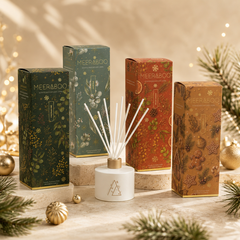 Four Meeraboo reed diffuser boxes in festive holiday packaging are arranged on stone blocks with a white reed diffuser bottle and reeds in front. The boxes are dark green, teal, rust red, and warm brown with gold-toned holiday illustrations, surrounded by pine branches, gold ornaments, and warm string lights.