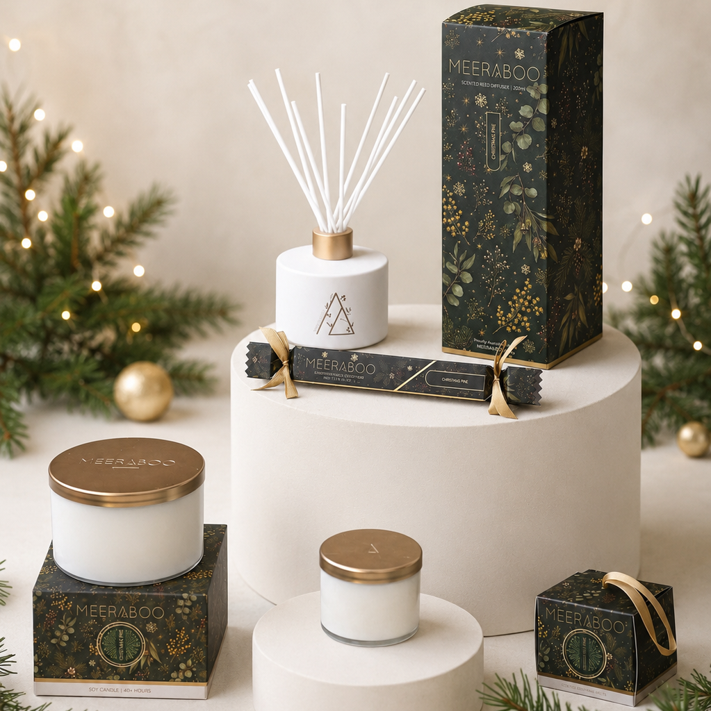 A styled product display of Meresbon scented candles and a reed diffuser set on white tiered pedestals, surrounded by pine branches, gold ornaments, and warm fairy lights.