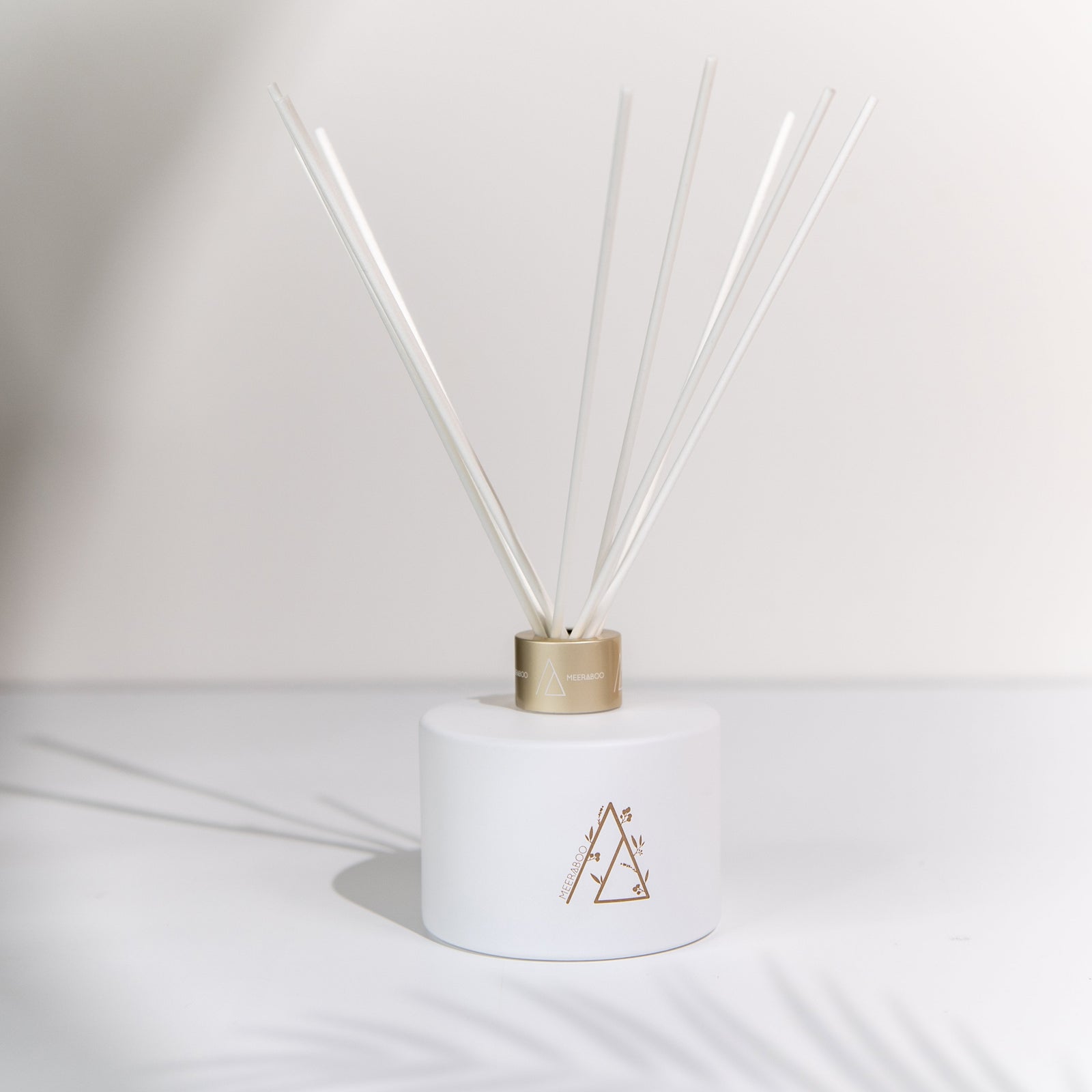 A white reed diffuser bottle with gold geometric detailing and white reeds, standing beside its white and gold branded box on a stone surface.