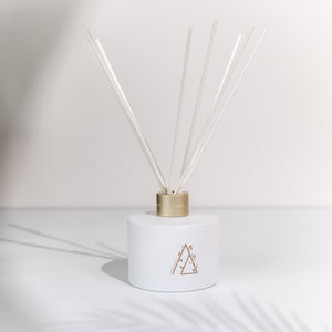 A white ceramic reed diffuser with a gold-toned metal collar and clear reeds, featuring a minimalist geometric logo on the front, set against a white background with soft palm leaf shadows.