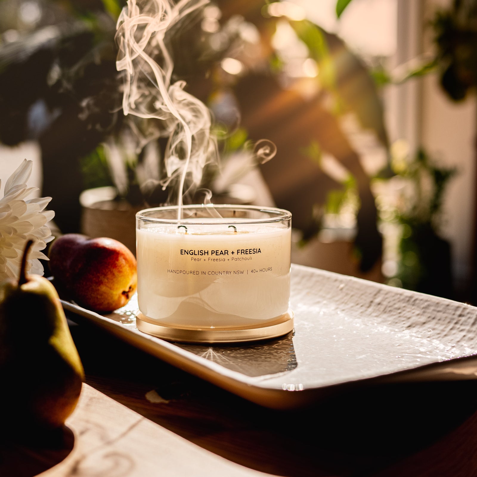 A lit double-wick candle in a clear glass jar sits on a white tray on a wooden table, styled with pears, white flowers, and a woven bag in a warm indoor setting.