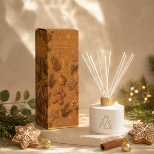 A festive Christmas-themed reed diffuser set by Meeraboo, featuring a white cylindrical diffuser with white reeds and a matching brown patterned box decorated with gingerbread men, pinecones, and snowflakes. The set is styled on a stone slab with holiday decorations including a cinnamon stick, star-shaped cookies, and soft warm lights in the background.