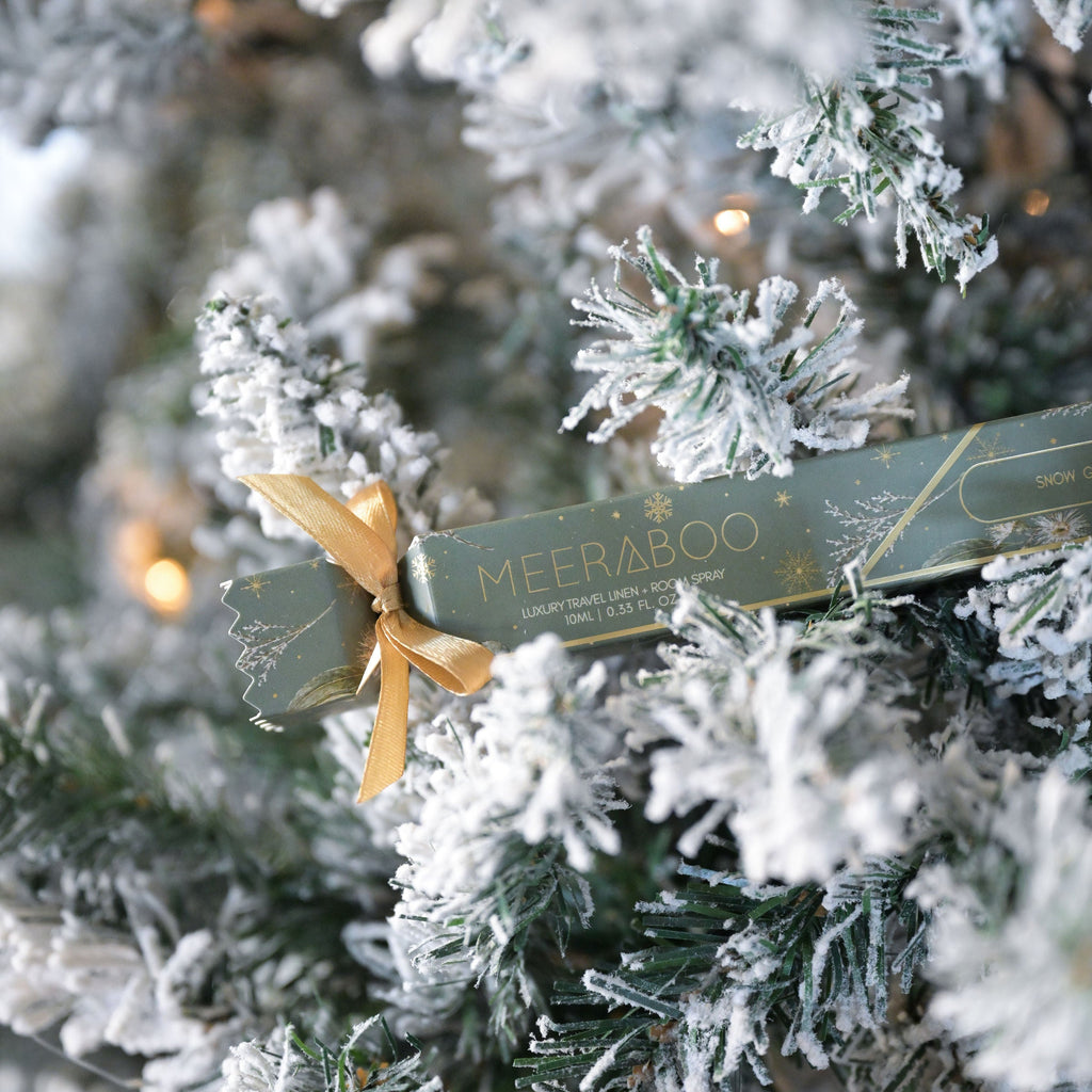 A close-up of a green MEERABOO gift box tied with a gold ribbon, nestled among frosted Christmas tree branches.