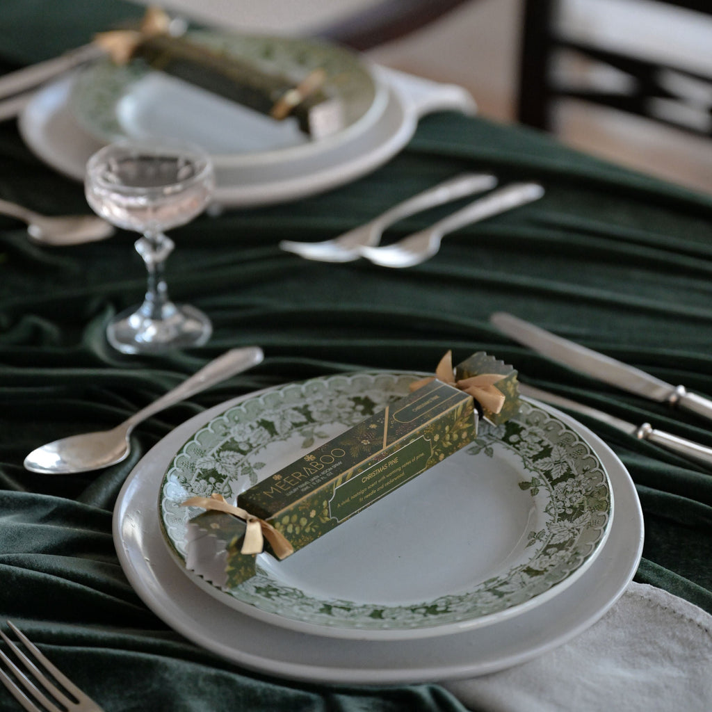 A styled dining table setting with white plates, silver cutlery, a small glass, and a dark green velvet tablecloth. A decorative green and gold box tied with a ribbon is placed on each plate, creating a festive table setting scene.