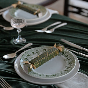 A styled dining table setting with white plates, silver cutlery, a small glass, and a dark green velvet tablecloth. A decorative green and gold box tied with a ribbon is placed on each plate, creating a festive table setting scene.