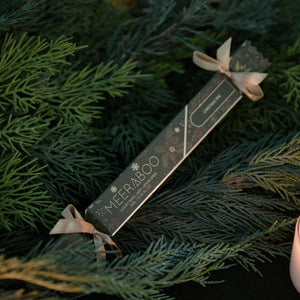 A packaged linen spray wrapped in a long dark gray paper sleeve with the brand name MEERABOO visible, tied with a small gold ribbon, resting among green evergreen branches with warm candlelight in the background.