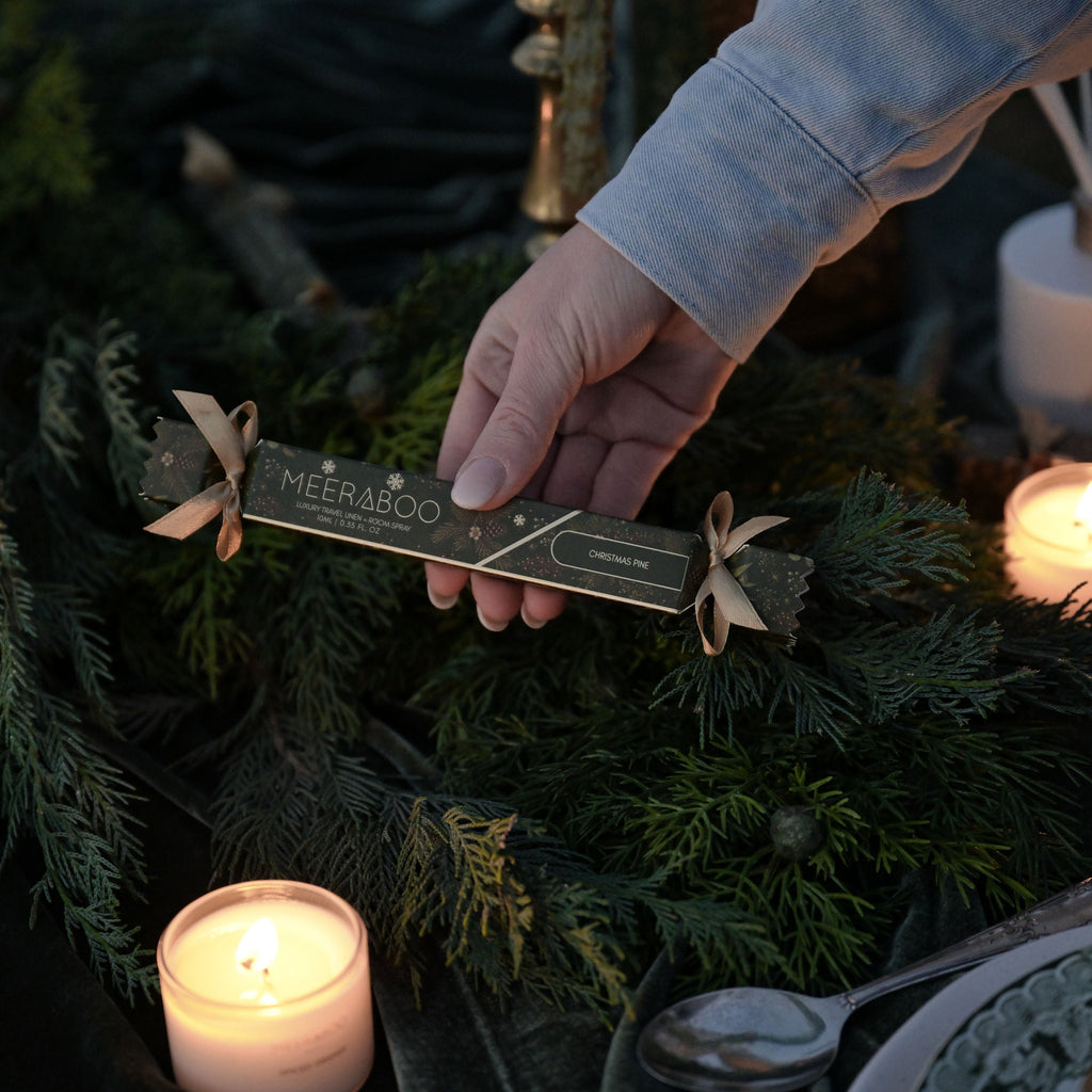 A hand holding a MEERABOO linen spray wrapped in dark paper, surrounded by green pine branches, lit candles, and a dark velvet cloth.
