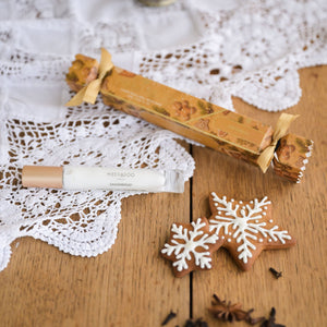 A wrapped Christmas cracker and a small linen spray rest on a white lace doily over a wooden table, with gingerbread cookies and star anise nearby.