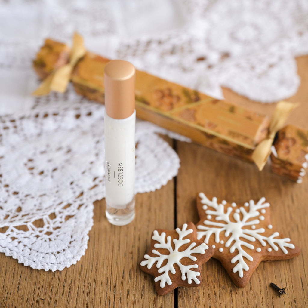A clear glass perfume bottle with a metallic rose gold cap stands on a wooden table next to a gingerbread cookie decorated with white icing snowflakes, a patterned gift box, and a white lace doily.