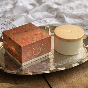 A Meeraboo candle shown with its decorative red and gold gift box and a white jar with a gold lid, styled on a silver tray over a wooden surface with a white lace cloth in the background.