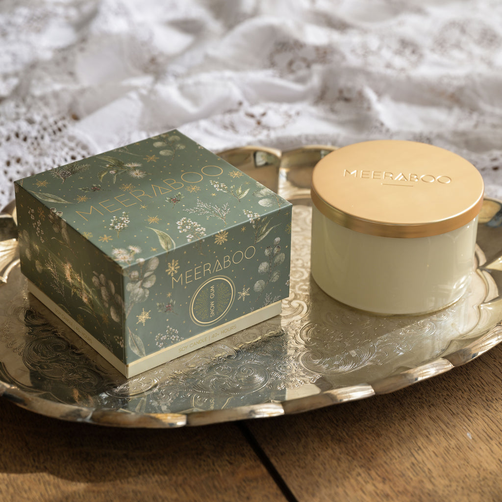 A boxed candle  and its cream jar sit on an ornate silver tray. The square box is muted teal-green with gold floral and star illustrations and the brand name MEERABOO visible on both the box and the gold-lidded jar.