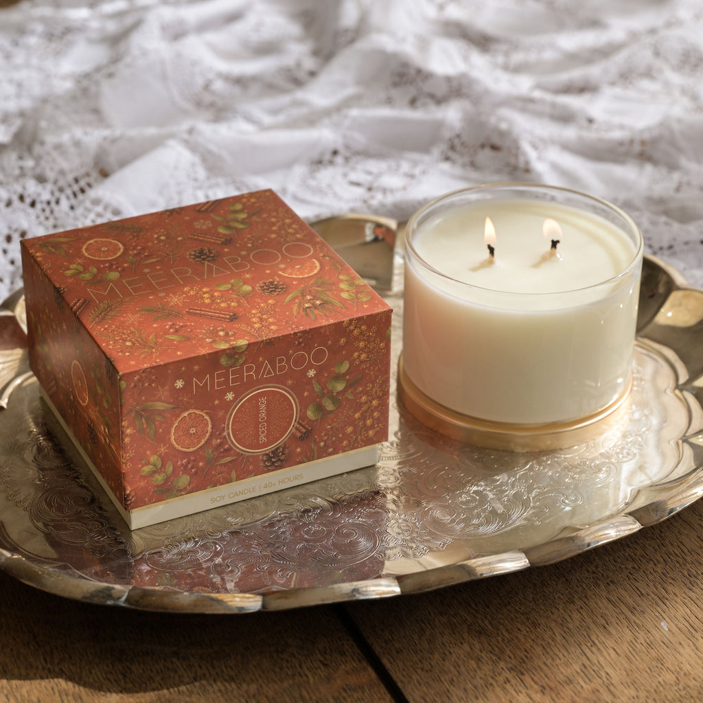A two-wick soy candle in a clear glass jar sits on a silver tray next to its decorative rust-orange patterned box, set on a wooden table with a white lace cloth in the background.