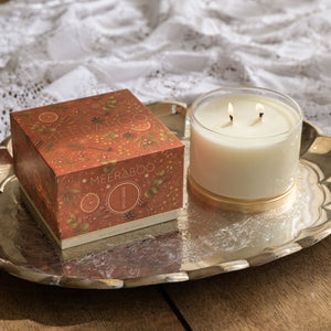 A two-wick soy candle in a clear glass jar sits on a silver tray next to its decorative rust-orange patterned box, set on a wooden table with a white lace cloth in the background.