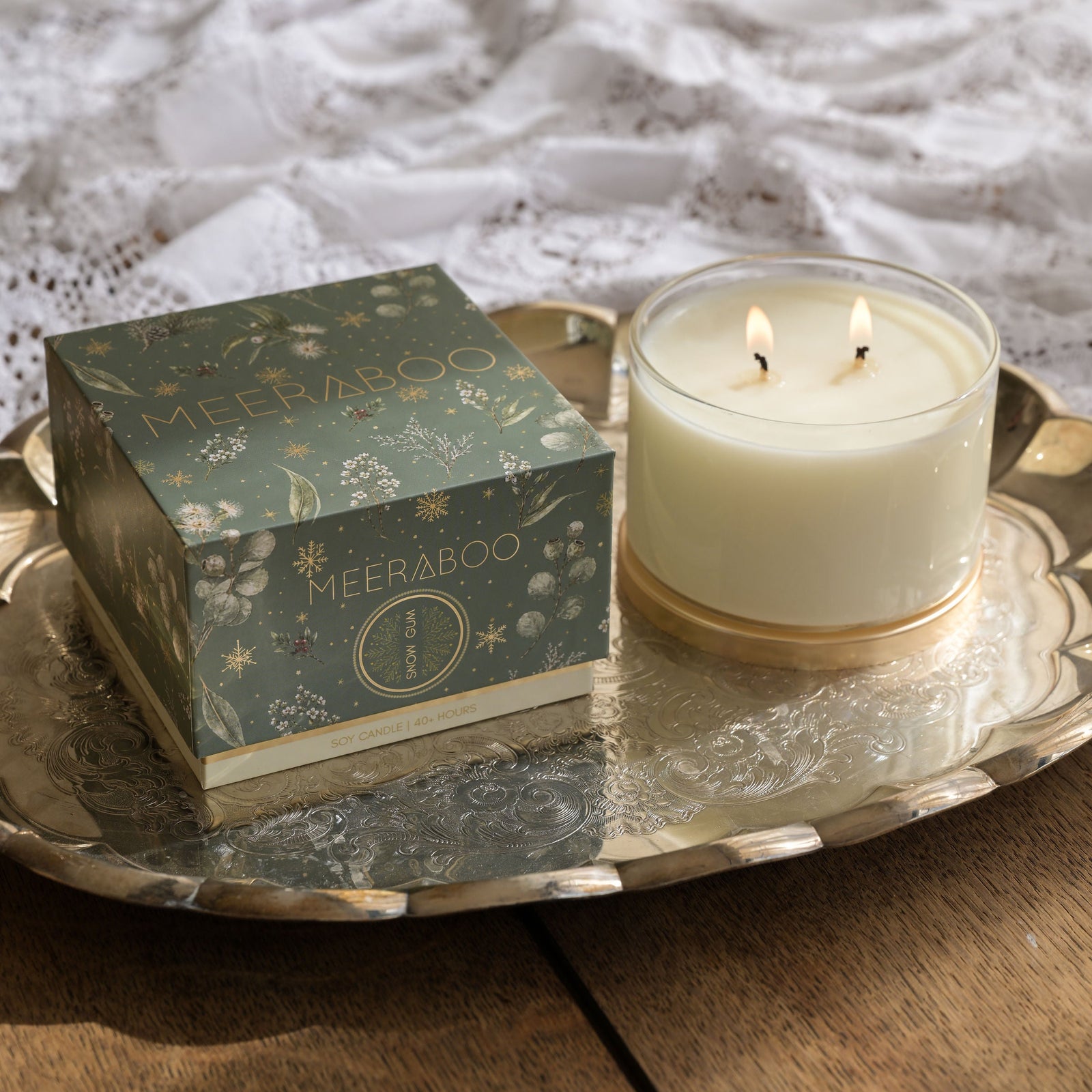 A lit white candle in a clear glass jar with a gold base sits beside its decorative teal and gold box. A hand holds a match above the candle.