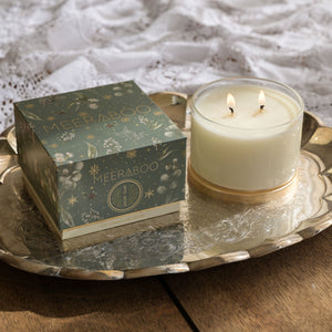A lit cream-colored candle in a clear glass jar sits on a silver tray next to its decorative green and gold box, labeled 'MEERABOO'. The tray is placed on a rustic wooden table with a white lace cloth in the background.