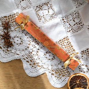 An orange Christmas cracker with gold foil details and ribbon bows, styled on a white lace doily with dried orange slices and star anise on a wooden surface.