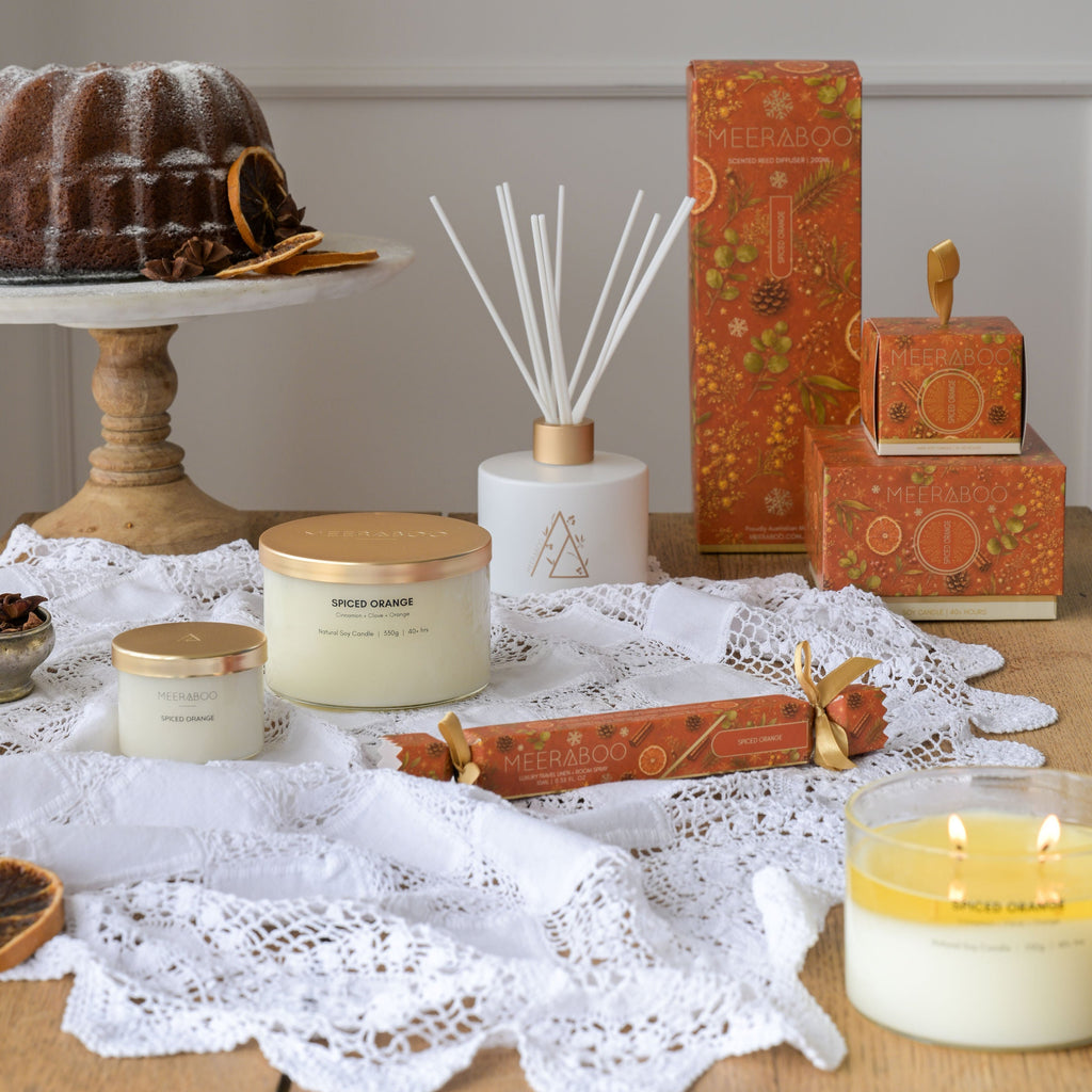 A styled tabletop arrangement of home fragrance products, including candles, a reed diffuser, and patterned boxes, set on white lace over a wooden table with a neutral wall behind.