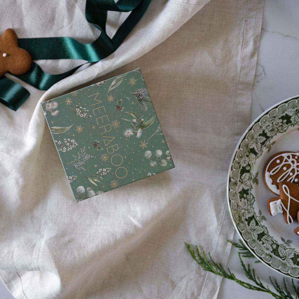 A square mint green gift box with gold foil 'MEERABOO' lettering and botanical illustrations, styled on a white linen cloth with a dark green ribbon, a gingerbread cookie, and a plate of decorated cookies on a white marble surface.