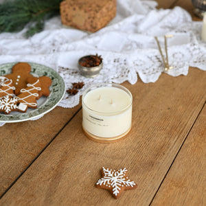 A lit cream-colored candle in a clear glass jar with a gold-toned base sits on a rustic wooden table, surrounded by holiday decorations including gingerbread cookies, a lace runner, pine branches, and a small bowl of spices.