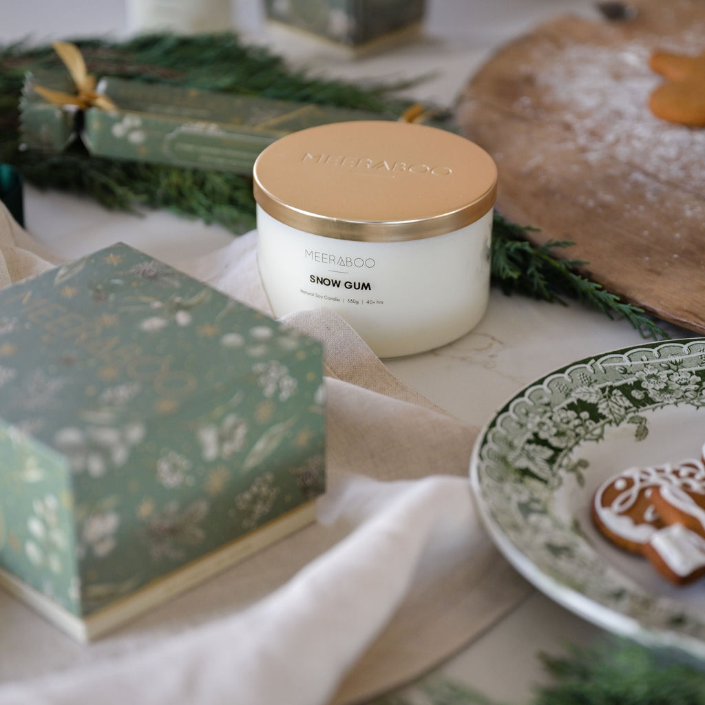 A candle jar labeled 'meeraboo' and 'SNOW GUM' sits on a styled tabletop with green pine branches, a patterned gift box, a plate with a gingerbread cookie, and soft neutral fabric in the background.