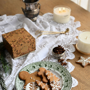 A styled holiday tabletop scene with a green patterned plate holding decorated gingerbread cookies, surrounded by white lace doilies, candles, a small box, and pine branches on a wooden table.