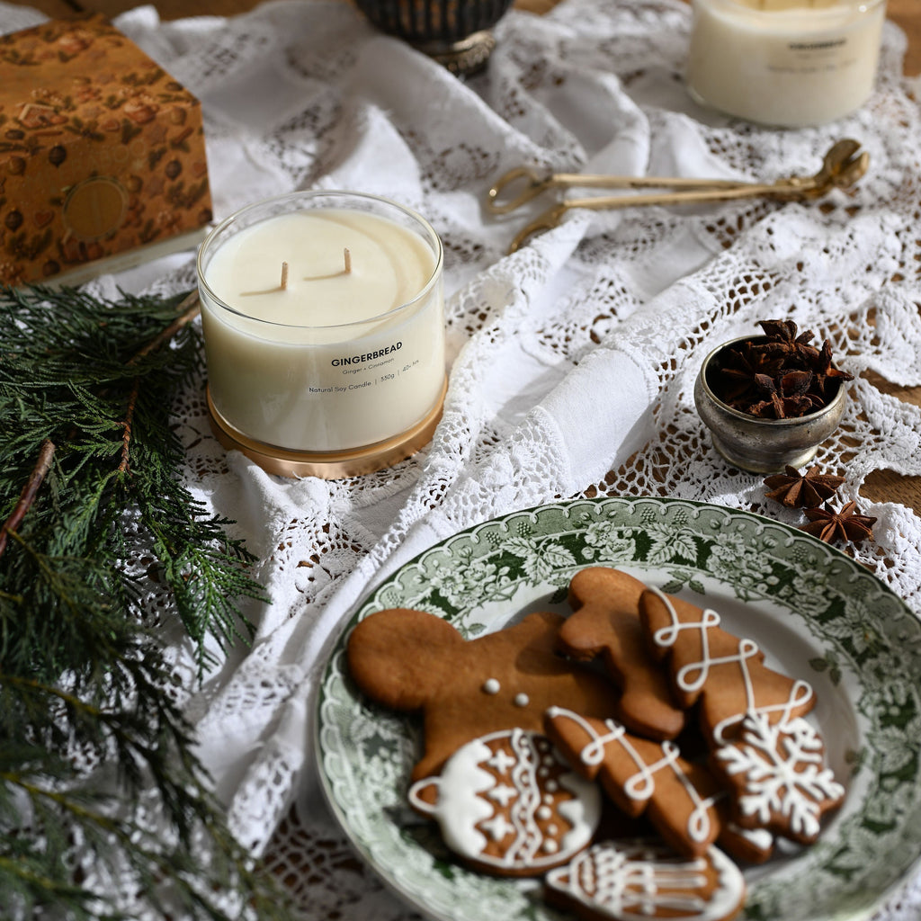 A styled holiday tabletop scene with three lit candles on white lace fabric, gingerbread cookies on a green patterned plate, evergreen branches, spices, and a wrapped gift box on a wooden surface.