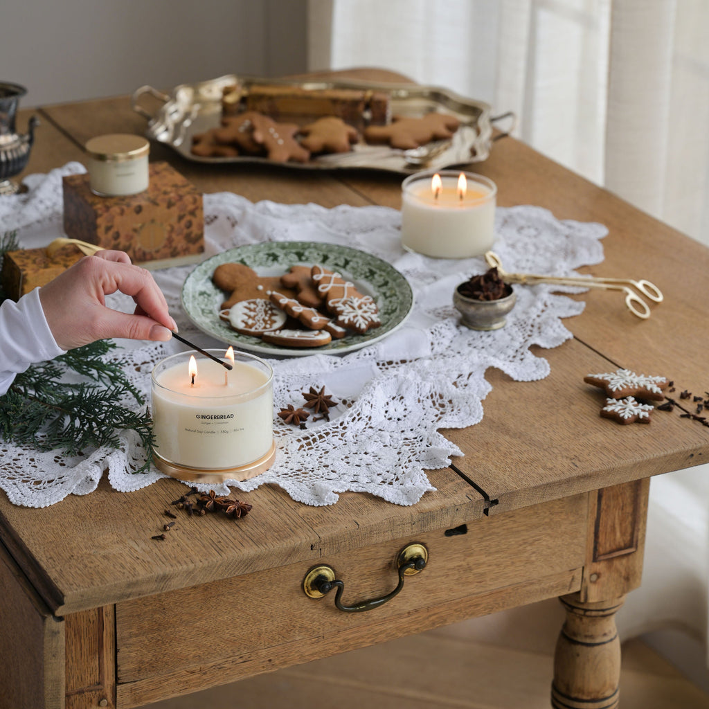A cozy tabletop scene with lit candles, gingerbread cookies on a plate, lace runner, and a hand lighting a candle near a wooden table in a softly lit room.