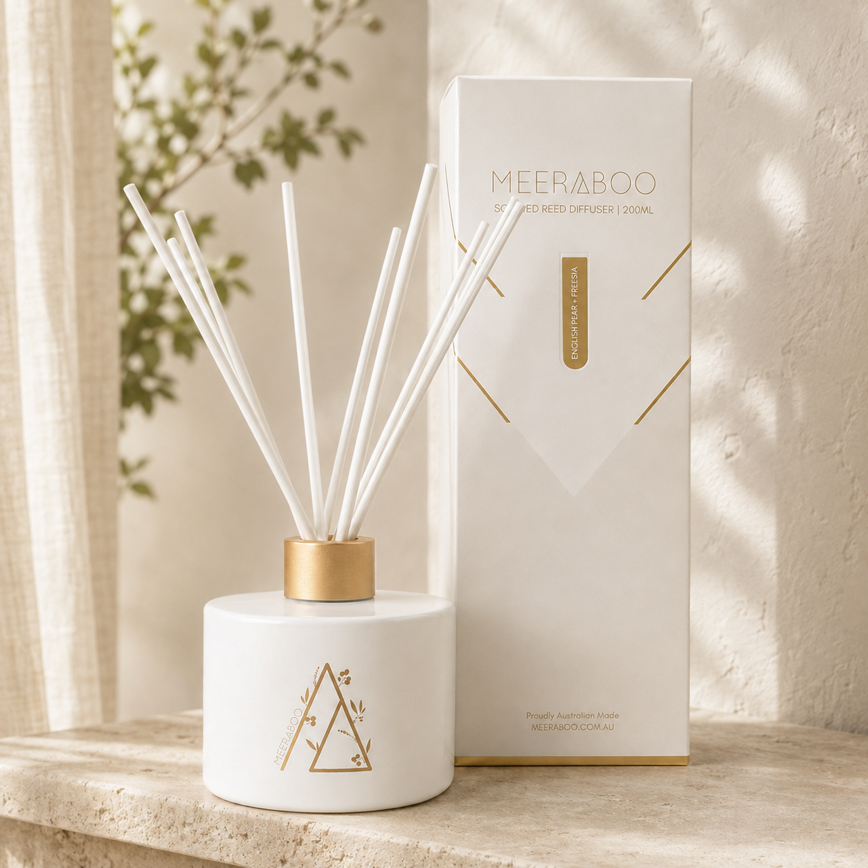 A white reed diffuser bottle with gold geometric detailing and white reeds, standing beside its white and gold branded box on a stone surface.