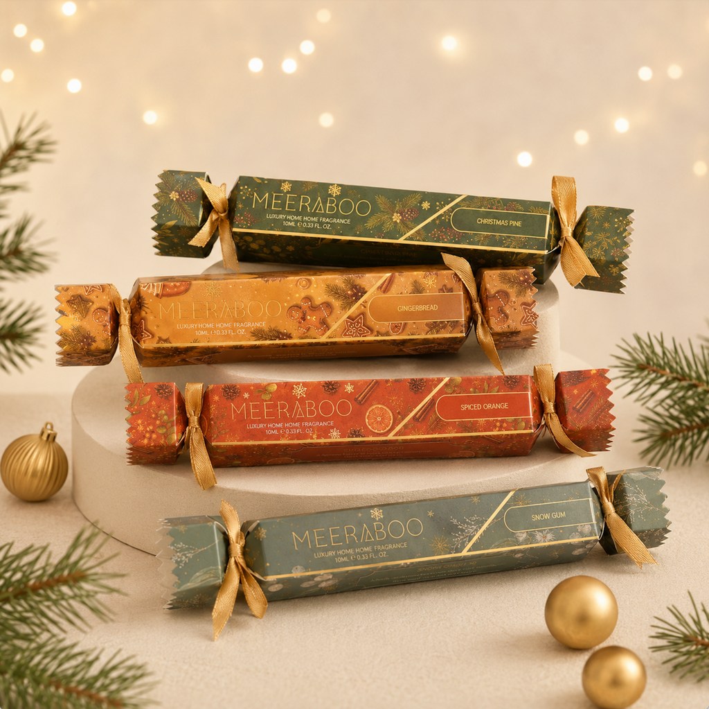 Four MEERABOO Christmas crackers arranged on a white pedestal with festive green and gold holiday decorations, including pine branches, gold ornaments, and warm string lights in the background.