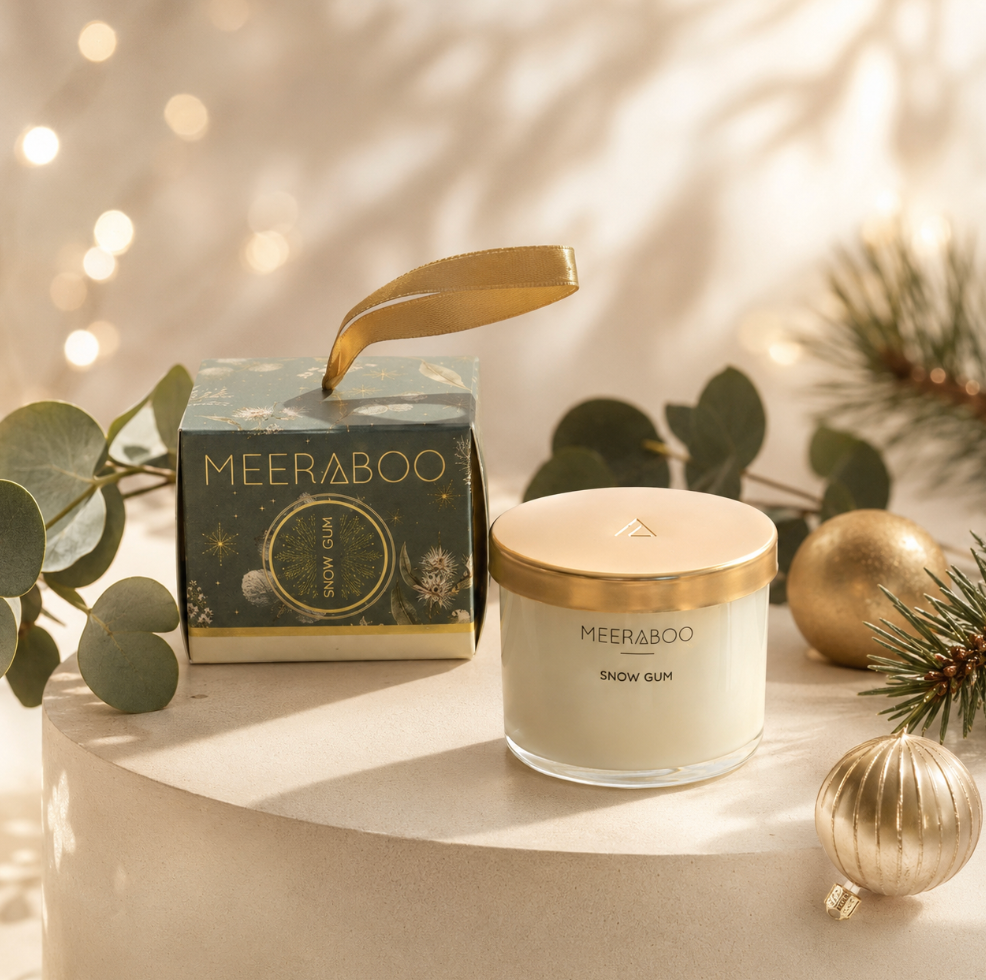 A MEERABOO Snow Gum candle in a clear glass jar with a gold lid sits beside its matching decorative box on a round pedestal, styled with eucalyptus leaves, pine branches, and gold ornaments in a warm holiday setting.
