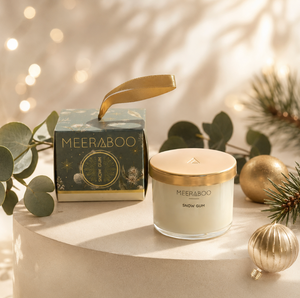 A MEERABOO Snow Gum candle in a clear glass jar with a gold lid sits beside its matching decorative box on a round pedestal, styled with eucalyptus leaves, pine branches, and gold ornaments in a warm holiday setting.