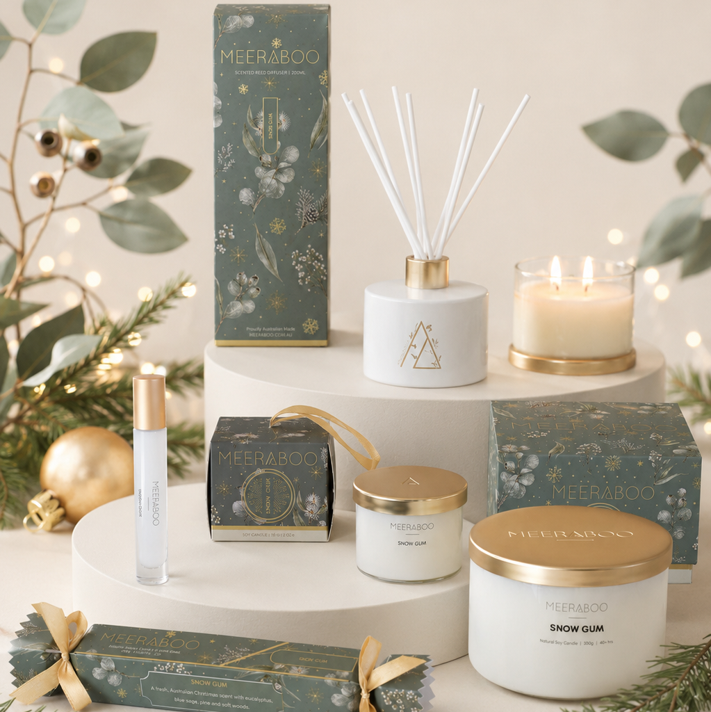 A styled product display of Meeraboo holiday home fragrance items, including a reed diffuser, candle, jar candle, roller bottle, and gift boxes, arranged on white pedestals with eucalyptus and gold ornaments.