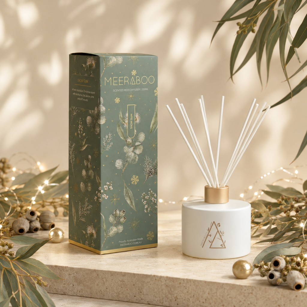 A reed diffuser bottle with white reeds and a gold cap stands beside its matching decorative box. The box is sage green with gold botanical illustrations and the brand name MEERABOO visible.