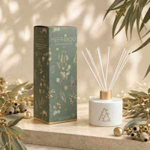 A reed diffuser bottle with white reeds and a gold cap stands beside its matching decorative box. The box is sage green with gold botanical illustrations and the brand name MEERABOO visible.
