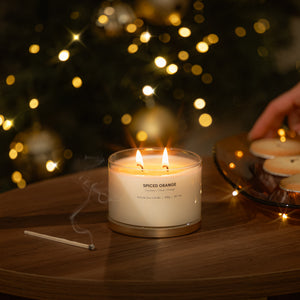 A lit double-wick candle labeled 'SPICED ORANGE' sits on a wooden table with a match nearby and a hand placing more candles on a plate in the background, set against a warm, festive bokeh of lights.
