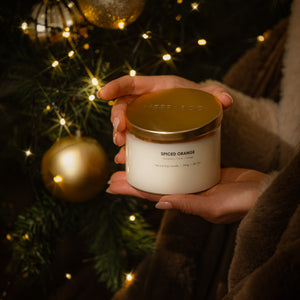 A person's hands holding a white candle jar with a gold lid, labeled 'SPICED ORANGE', set against a festive background of a Christmas tree with gold ornaments and warm string lights.