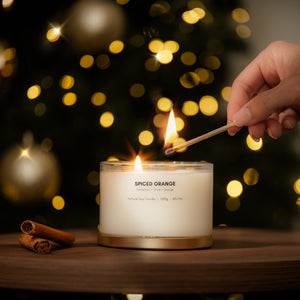 A hand lighting a candle with a match on a wooden table, with a blurred Christmas tree and warm bokeh lights in the background. The candle is in a clear glass jar with white wax and a gold-toned base, labeled 'SPICED ORANGE'.