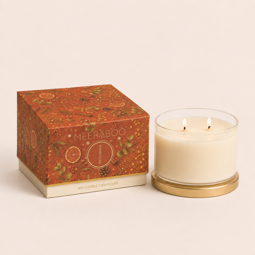 A lit double-wick soy candle in a clear glass jar with a gold-toned base, shown next to its decorative orange-brown patterned box. The box is branded MEERABOO and reads 'Scent of the Season'.