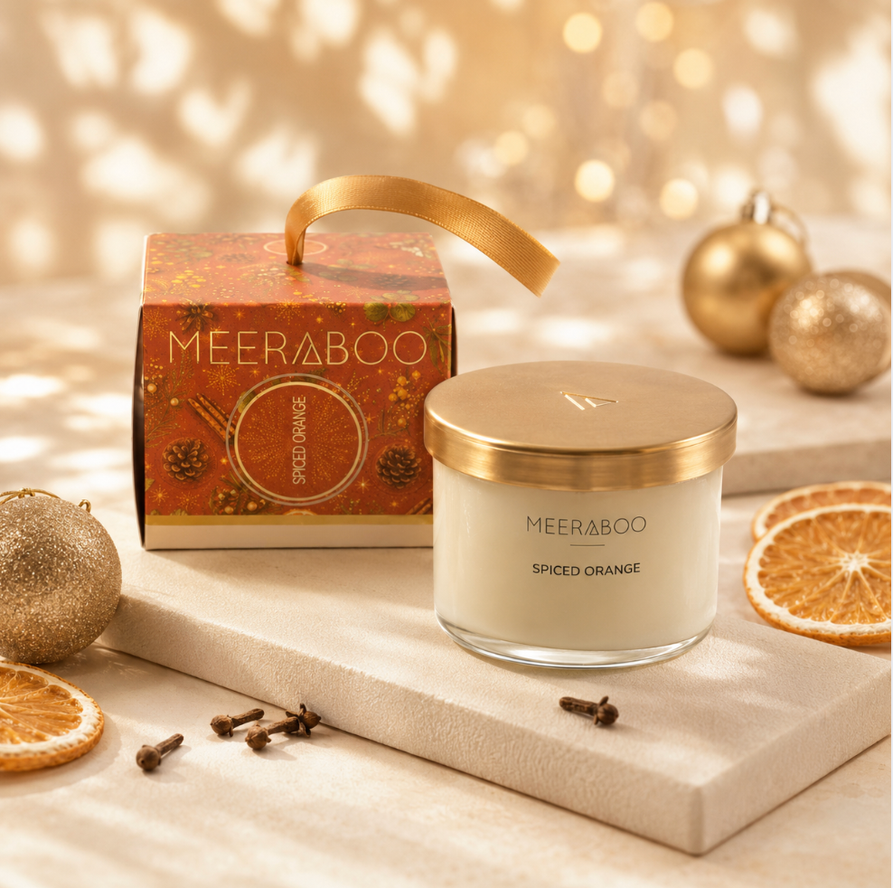 A MEERABoo Spiced Orange scented candle in a clear glass jar with a gold lid, styled on a beige stone slab with its matching orange-patterned box, gold ornaments, dried orange slices, and cloves against a warm bokeh holiday background.