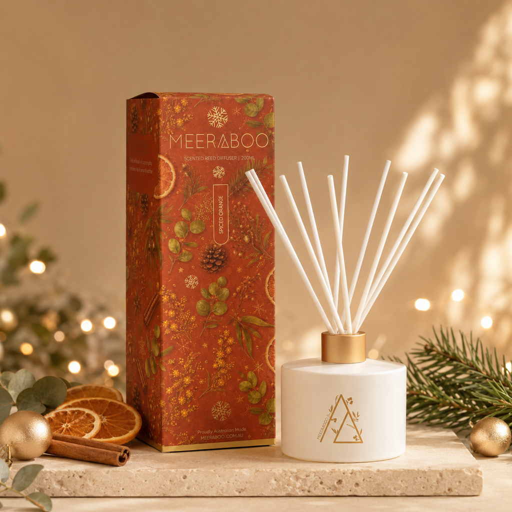A reed diffuser set with a white ceramic bottle, gold cap, and white reeds, displayed next to its orange patterned retail box on a stone surface with holiday decor and warm string lights in the background.