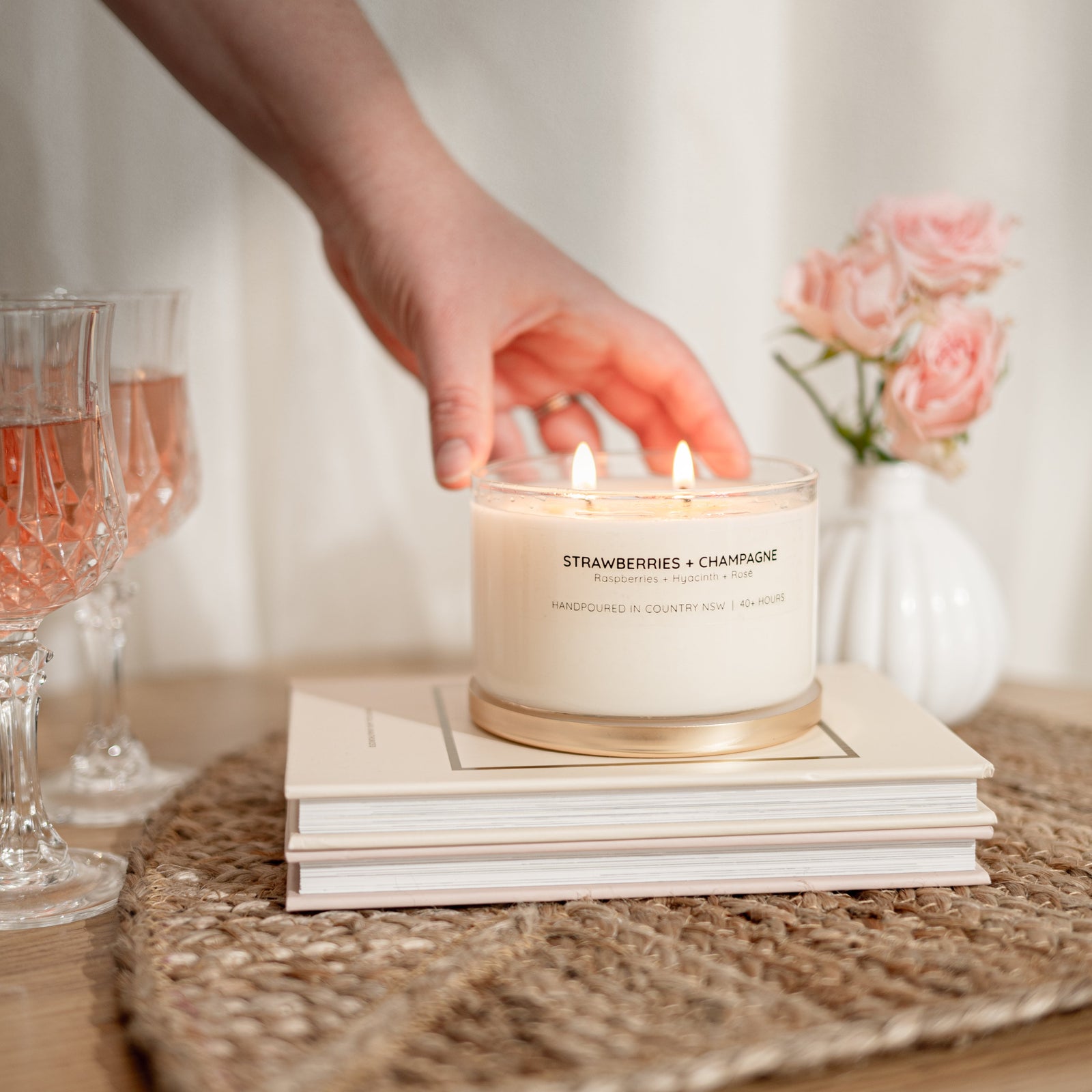 A lit candle in a clear glass jar sits on stacked books on a woven placemat, with a person's hand reaching toward it. Pink champagne flutes and pink roses in a small white vase are blurred in the background.