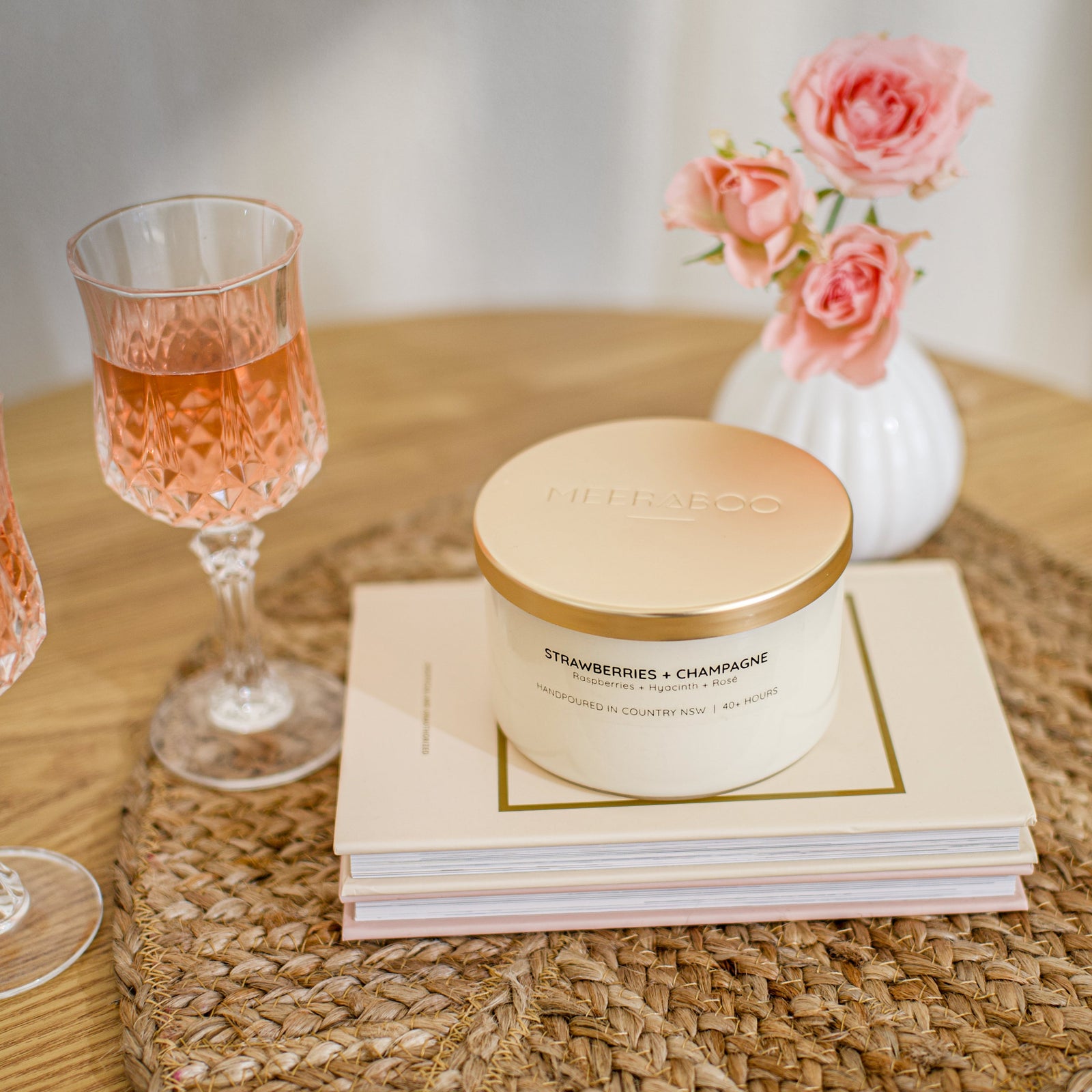 A lit candle in a clear glass jar sits on stacked books on a woven placemat, with a person's hand reaching toward it. Pink champagne flutes and pink roses in a small white vase are blurred in the background.
