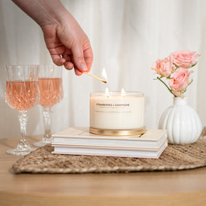 A lit scented candle in a clear glass jar with a gold-toned base sits on stacked books on a woven placemat, with a hand holding a match above it. Two pink drinks in glasses and a white vase of pink roses are nearby against a soft cream curtain background.